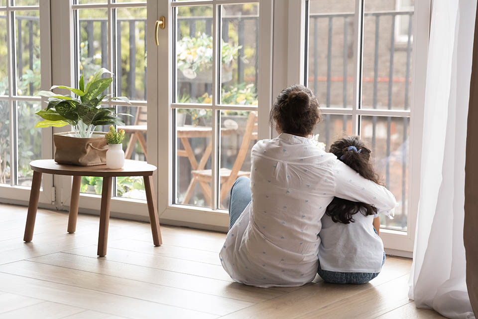 Rückansicht einer schwarzhaarigen jungen Mutter und eines kleinen Tochtermädchens, die zu Hause auf dem Boden sitzen, sich liebevoll und fürsorglich umarmen und auf das Panoramafenster und den gemütlichen Balkon blicken. Mutterschaft, Immobilienkonzept